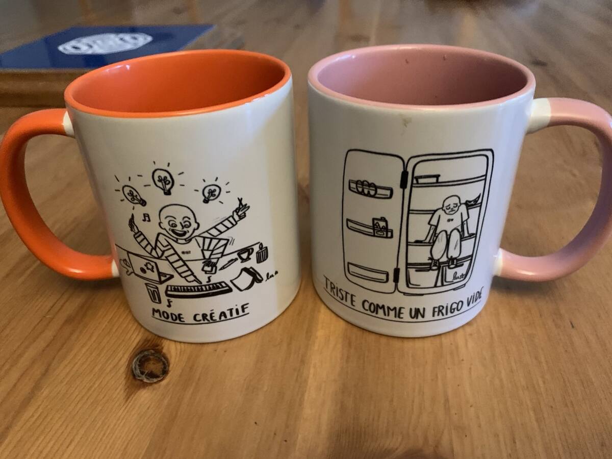 Deux tasses posées sur une table en bois : l'une, orange, illustrée d'un personnage débordant d'idées et titrée « Mode créatif » ; l'autre, rose, montrant un personnage assis dans un réfrigérateur ouvert, titrée « Triste comme un frigo vide ».